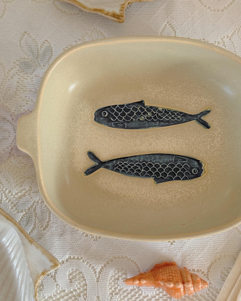 May include: Cream-colored ceramic serving dish with two dark blue fish designs. The oval dish has small handles on each side. A seashell is visible in the bottom right corner. The dish is on a white lace tablecloth.