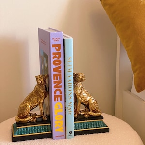 K&ouml;nnte beinhalten: Goldfarbene Geparden-Buchst&uuml;tzen mit schwarzem und t&uuml;rkisem Sockel, die zwei B&uuml;cher halten. Die B&uuml;cher tragen die Titel "Provence Glory" und "Slim Aarons Style". Die Buchst&uuml;tzen stehen auf einer wei&szlig;en, strukturierten Oberfl&auml;che.