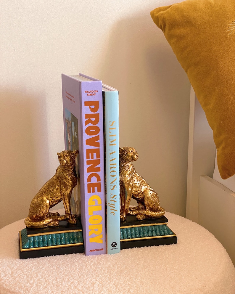 K&ouml;nnte beinhalten: Goldfarbene Geparden-Buchst&uuml;tzen mit schwarzem und t&uuml;rkisem Sockel, die zwei B&uuml;cher halten. Die B&uuml;cher tragen die Titel "Provence Glory" und "Slim Aarons Style". Die Buchst&uuml;tzen stehen auf einer wei&szlig;en, strukturierten Oberfl&auml;che.