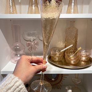 May include: A tall, champagne-colored champagne flute with a textured surface is held in front of a shelf filled with glassware. Other items include small glasses, a decorative bottle, and a tray with drink stirrers. The overall theme is elegant barware.