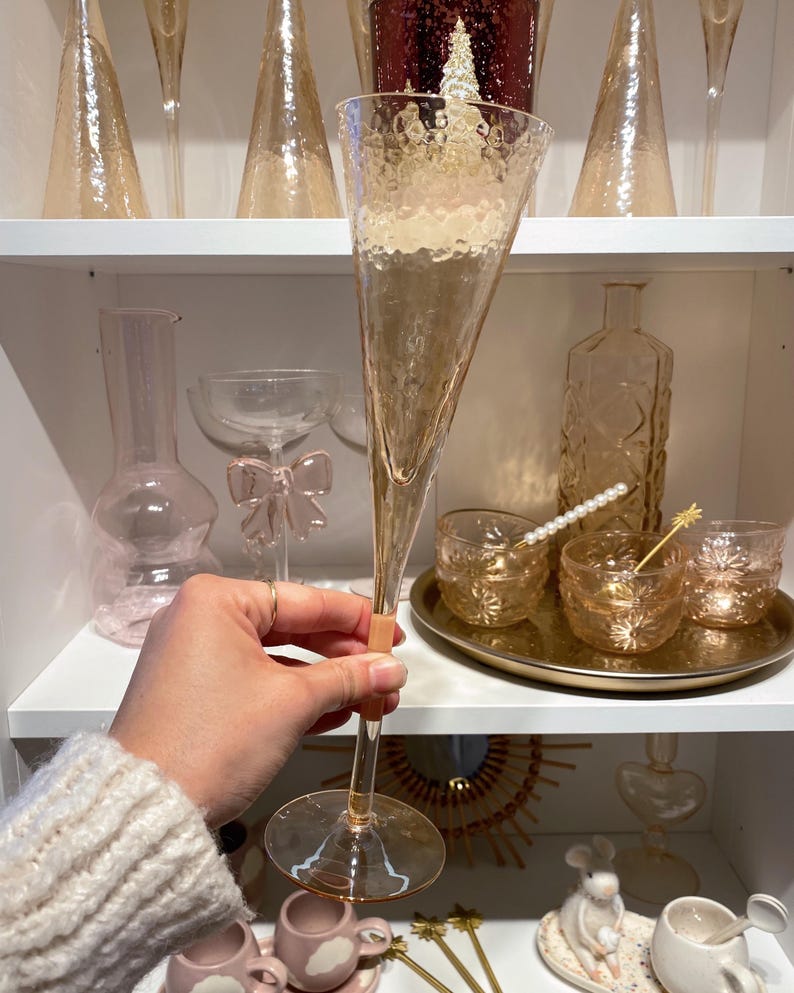 May include: A tall, champagne-colored champagne flute with a textured surface is held in front of a shelf filled with glassware. Other items include small glasses, a decorative bottle, and a tray with drink stirrers. The overall theme is elegant barware.