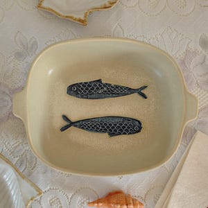 May include: A beige ceramic serving dish with two stylized blue fish designs. The dish has a rectangular shape with rounded corners and small handles. A seashell and a napkin are also visible.