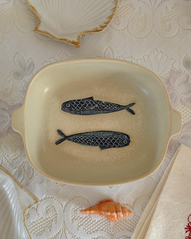 May include: A beige ceramic serving dish with two stylized blue fish designs. The dish has a rectangular shape with rounded corners and small handles. A seashell and a napkin are also visible.