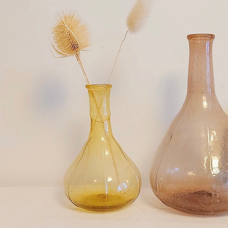May include: Two glass vases, one amber and one clear, with a dried flower arrangement. The amber vase is round and has a bulbous shape. The clear vase is taller and has a more traditional shape.