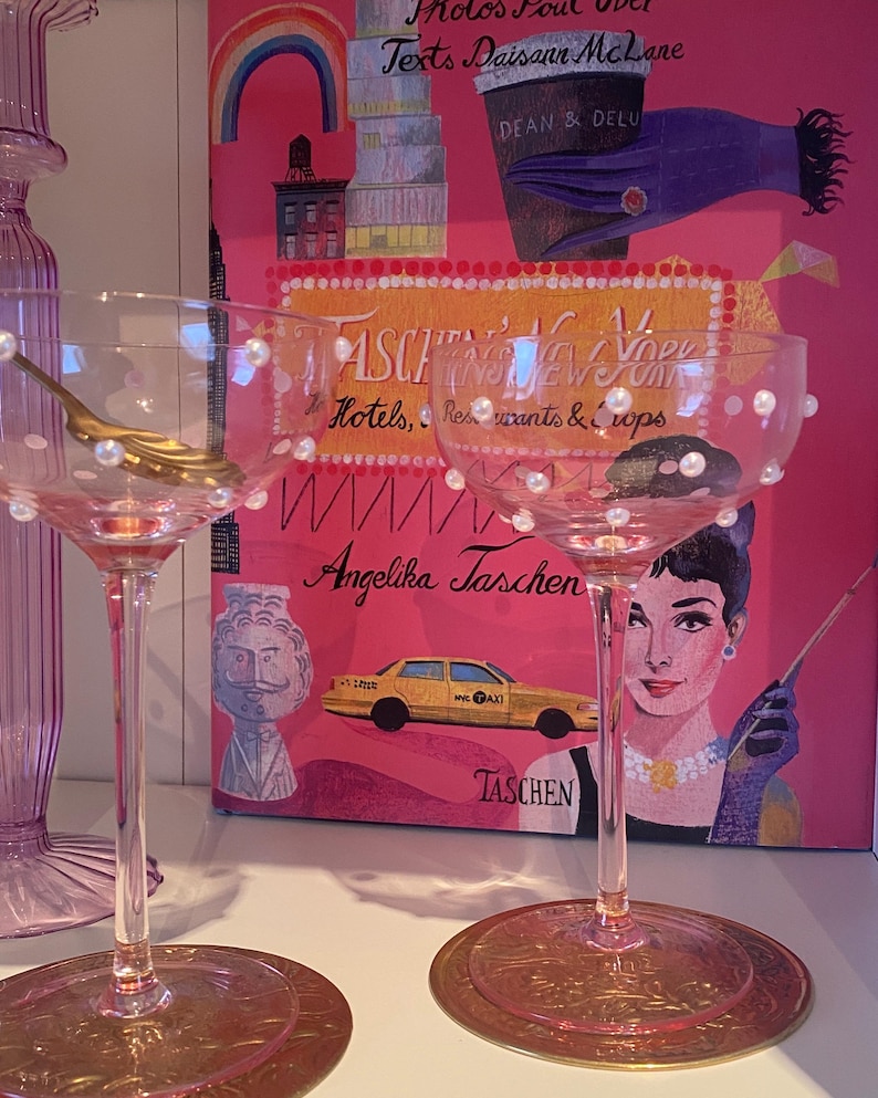 May include: Two pink champagne glasses with pearl accents and gold spoons. The glasses sit on gold coasters. A pink book cover with the text "TASCHEN'S NEW YORK" and an illustration of Audrey Hepburn is in the background.