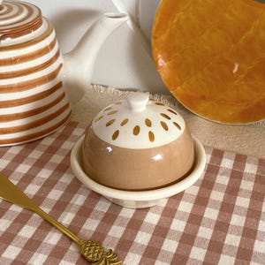 Könnte beinhalten: Eine Keramik-Butterdose mit braunem Boden und weißem Deckel mit goldenen Akzenten. Die Dose steht auf einem weißen Sockel, und ein goldfarbener Streuer mit Ananas-Design befindet sich im Vordergrund. Eine gestreifte Teekanne ist im Hintergrund.