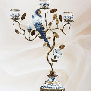 Puede incluir: Un candelero de metal dorado con un pájaro de porcelana azul y blanco y tres portavelas. La base del candelero también está decorada con porcelana azul y blanco.