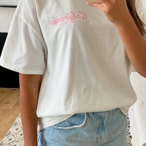 K&ouml;nnte beinhalten: Ein wei&szlig;es T-Shirt mit einem rosafarbenen Aufdruck, auf dem "Dreaming California" steht. Das Shirt ist oversized und hat eine lockere Passform. Das Model tr&auml;gt das Shirt zu hellblauen Jeans-Shorts.