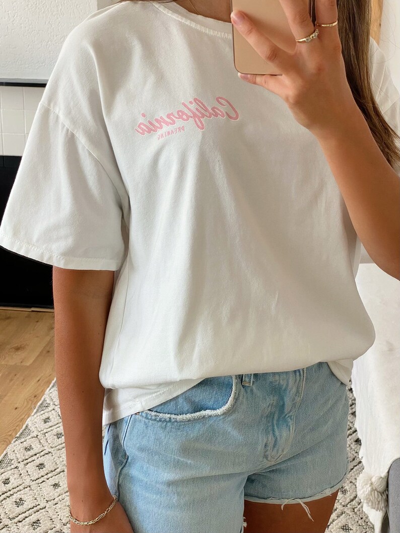 K&ouml;nnte beinhalten: Ein wei&szlig;es T-Shirt mit einem rosafarbenen Aufdruck, auf dem "Dreaming California" steht. Das Shirt ist oversized und hat eine lockere Passform. Das Model tr&auml;gt das Shirt zu hellblauen Jeans-Shorts.