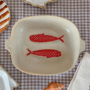 May include: A cream-colored ceramic serving dish with two red fish illustrations. The dish has a rectangular shape with rounded corners and small handles. Other seashell-themed decorative items are also visible.