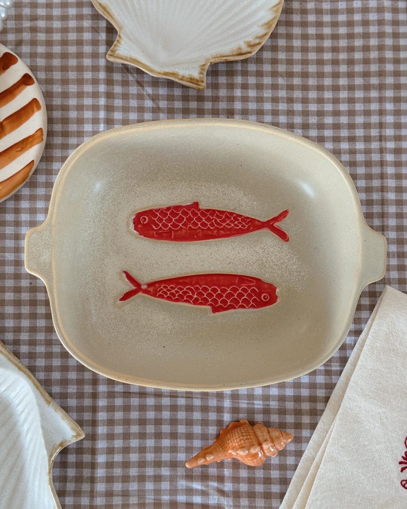 May include: A cream-colored ceramic serving dish with two red fish illustrations. The dish has a rectangular shape with rounded corners and small handles. Other seashell-themed decorative items are also visible.