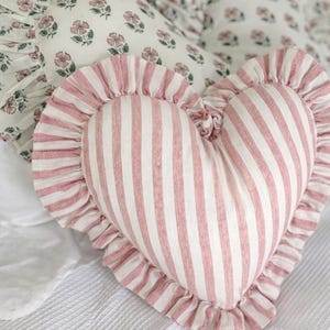 Può includere: Un cuscino a forma di cuore con strisce verticali rosa e bianche e un bordo a balze. Altri cuscini con motivi floreali sono sullo sfondo. I cuscini sono su un copriletto bianco, creando un'estetica accogliente e romantica.