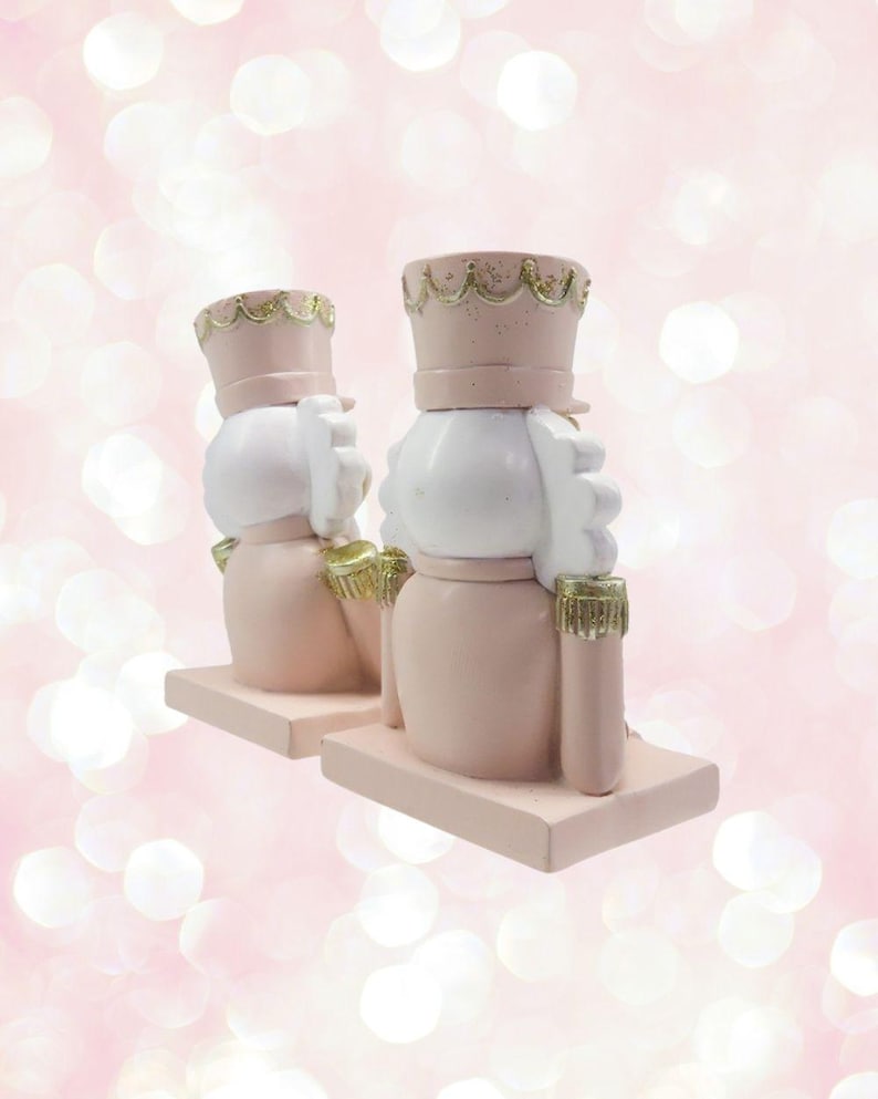 May include: A pair of pink and white nutcracker bookends with gold accents. The nutcrackers are wearing hats and have a whimsical design.