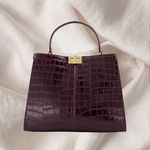 May include: A burgundy handbag with a crocodile-skin texture. The structured bag has a top handle and a gold-coloured clasp. The bag is set against a cream-coloured, textured background.