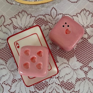 Könnte beinhalten: Ein rosa Keramik-Salz- und Pfefferstreuer-Set. Der Salzstreuer ist quadratisch mit herzförmigen Vertiefungen. Der Pfefferstreuer hat drei Löcher und ein Herz. Eine Spielkarte mit einem Herz-Ass ist ebenfalls im Bild.