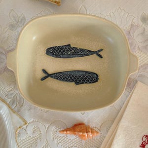 May include: A beige ceramic serving dish with two dark blue fish designs. The dish has a rectangular shape with rounded corners and small handles. A small orange seashell and a folded white napkin are also visible.