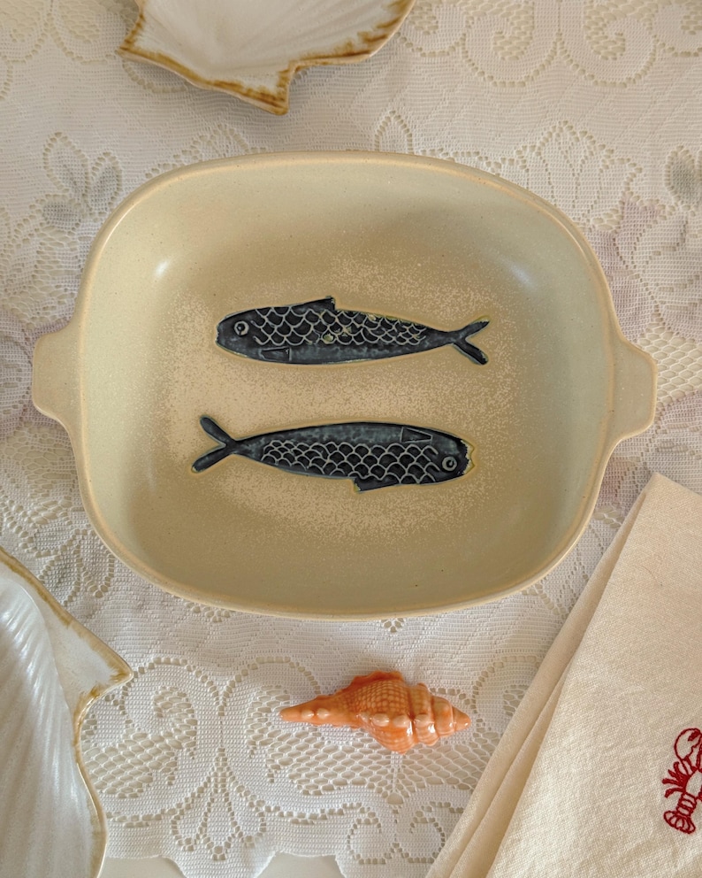 May include: A beige ceramic serving dish with two dark blue fish designs. The dish has a rectangular shape with rounded corners and small handles. A small orange seashell and a folded white napkin are also visible.