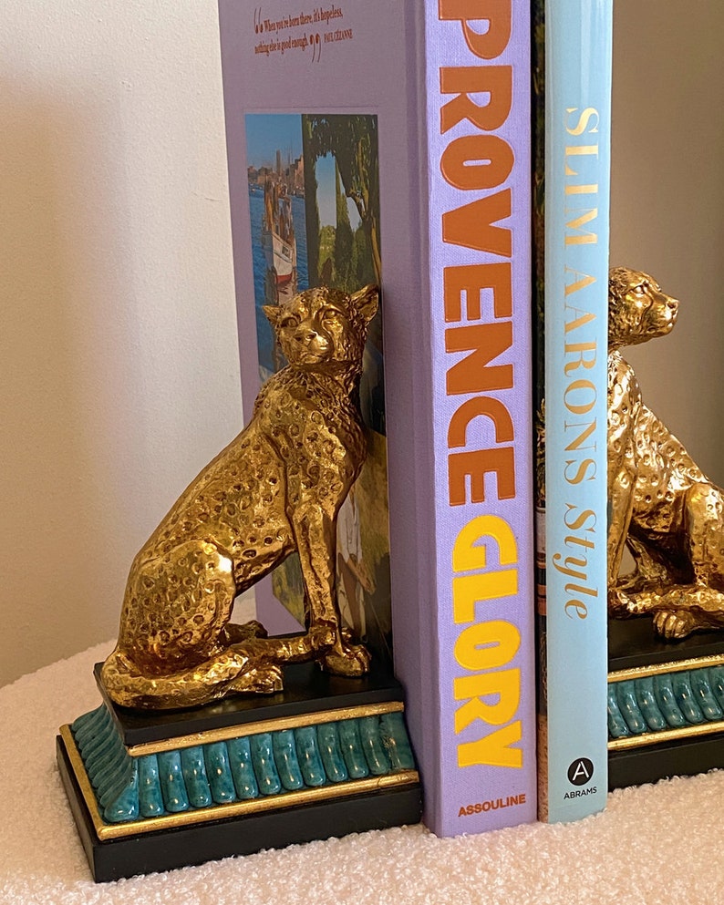 K&ouml;nnte beinhalten: Goldfarbene Geparden-Buchst&uuml;tzen mit schwarzen und t&uuml;rkisfarbenen Sockeln flankieren zwei B&uuml;cher. Die Buchst&uuml;tzen sind detailliert mit Flecken und einer sitzenden Pose. Die B&uuml;cher tragen die Titel "PROVENCE GLORY" und "SLIM AARONS Style".