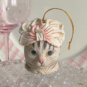 May include: A white ceramic cat ornament with a pink bow on its head. The ornament has a gold string for hanging.