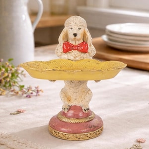 May include: A decorative poodle figurine with a red bow tie, holding a yellow tray. The base is pink with gold trim. The figurine is on a table with a floral arrangement and a white pitcher in the background.