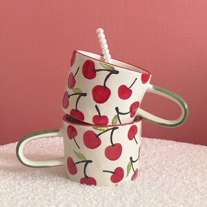 May include: Two white ceramic mugs with green handles and a red cherry pattern. The mugs are stacked on top of each other.