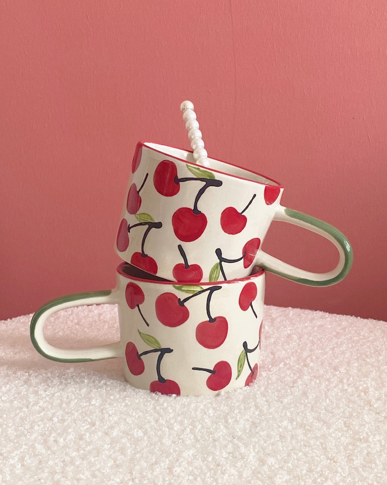 May include: Two white ceramic mugs with green handles and a red cherry pattern. The mugs are stacked on top of each other.