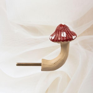 May include: A decorative mushroom-shaped cabinet knob. The knob has a red cap with white spots and a beige stem. The stem curves to meet a threaded metal rod for fixing. The design evokes a fairytale aesthetic.