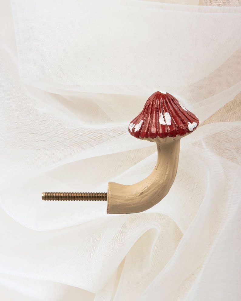 May include: A decorative mushroom-shaped cabinet knob. The knob has a red cap with white spots and a beige stem. The stem curves to meet a threaded metal rod for fixing. The design evokes a fairytale aesthetic.