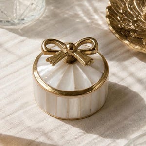 Puede incluir: Un joyero redondo con incrustaciones de n&aacute;car blanco, paneles verticales en los laterales y un patr&oacute;n de rayos de sol en la tapa. La caja est&aacute; decorada con un borde de metal dorado y un asa decorativa en forma de lazo de metal dorado.