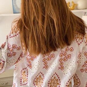 K&ouml;nnte beinhalten: Wei&szlig;e Baumwollbluse mit einem floralen Stickmuster in Rosa-, Orange- und Wei&szlig;t&ouml;nen. Die Stickerei zeigt ein sich wiederholendes Muster aus Blumen und Bl&auml;ttern.
