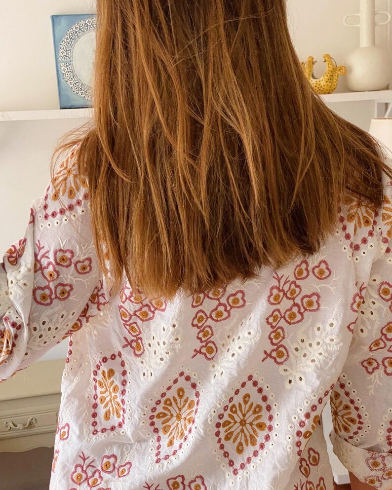 K&ouml;nnte beinhalten: Wei&szlig;e Baumwollbluse mit einem floralen Stickmuster in Rosa-, Orange- und Wei&szlig;t&ouml;nen. Die Stickerei zeigt ein sich wiederholendes Muster aus Blumen und Bl&auml;ttern.