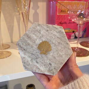 May include: A hexagonal marble coaster with a gold seashell emblem. The coaster has a gray and white speckled pattern. The gold seashell is centered on the coaster. The coaster is held in a person's hand.