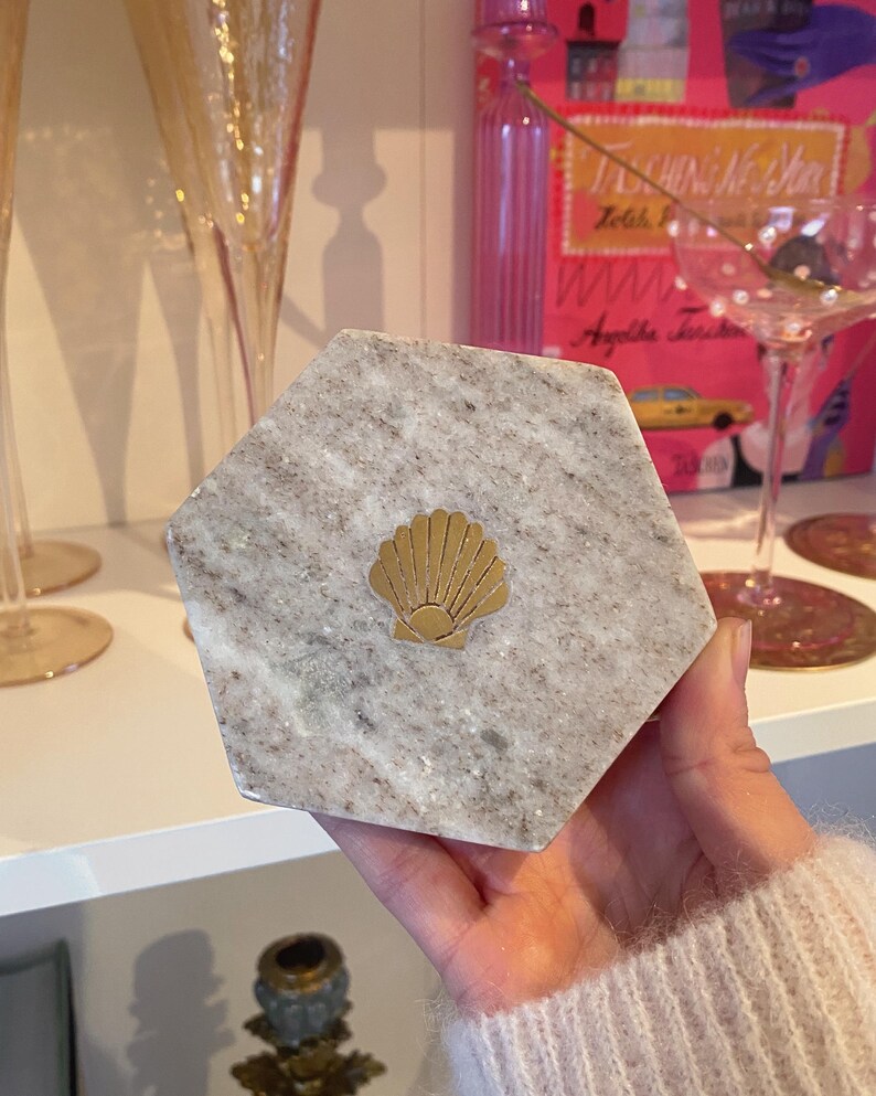May include: A hexagonal marble coaster with a gold seashell emblem. The coaster has a gray and white speckled pattern. The gold seashell is centered on the coaster. The coaster is held in a person's hand.