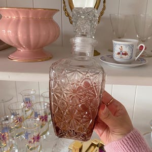 May include: A clear glass decanter with a geometric pattern and a crystal stopper. The bottom of the decanter has a gradient of amber color. The decanter is held in front of a shelf with other glassware and a pink vase.