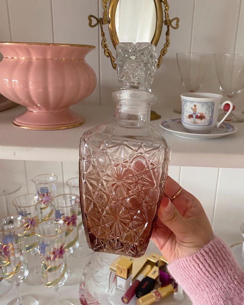 May include: A clear glass decanter with a geometric pattern and a crystal stopper. The bottom of the decanter has a gradient of amber color. The decanter is held in front of a shelf with other glassware and a pink vase.
