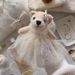 May include: A plush teddy bear wearing a cream-colored tulle dress with sparkling accents and a bow. The bear has a black nose and small eyes. Other items include a white handbag with a teddy bear applique, a white sweater, and jewelry.