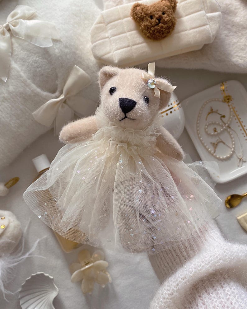 May include: A plush teddy bear wearing a cream-colored tulle dress with sparkling accents and a bow. The bear has a black nose and small eyes. Other items include a white handbag with a teddy bear applique, a white sweater, and jewelry.