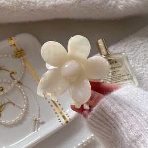 May include: A pearlescent white flower-shaped hair claw clip. The clip has six petals and a rounded center. The clip is held in a hand, with a white sweater sleeve visible. Other accessories are in the background.