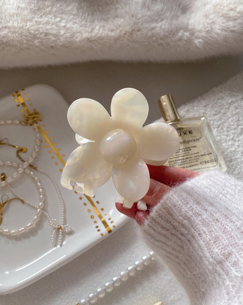 May include: A pearlescent white flower-shaped hair claw clip. The clip has six petals and a rounded center. The clip is held in a hand, with a white sweater sleeve visible. Other accessories are in the background.
