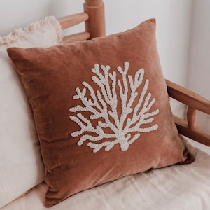 Pode incluir: Uma almofada de veludo marrom com um design de coral branco perolado. A almofada está sentada em um sofá de linho branco.