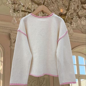 May include: A white sweater with a pink trim around the neckline, sleeves, and hem.