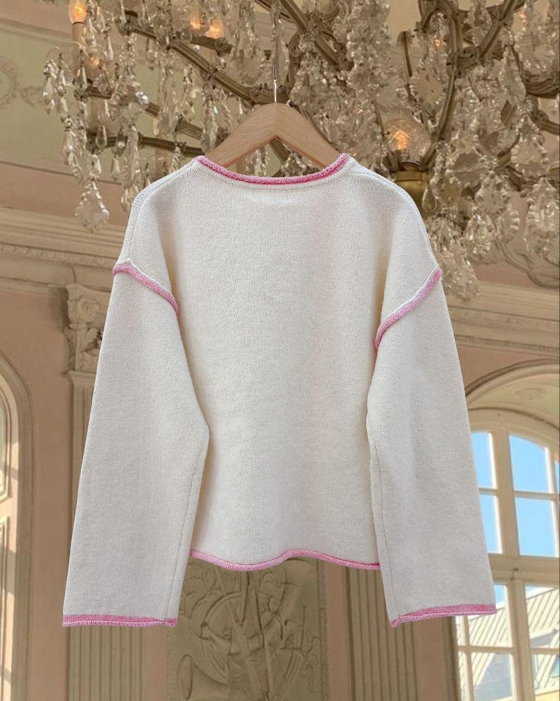 May include: A white sweater with a pink trim around the neckline, sleeves, and hem.
