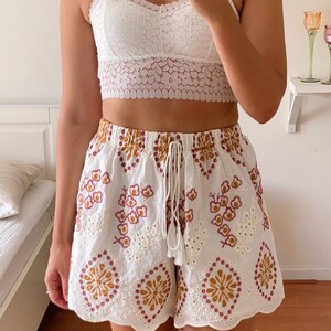 K&ouml;nnte beinhalten: Wei&szlig;e H&auml;kelshorts mit einem gewellten Saum und gestickten Blumen- und geometrischen Mustern in Braun-, Rosa- und Wei&szlig;t&ouml;nen.