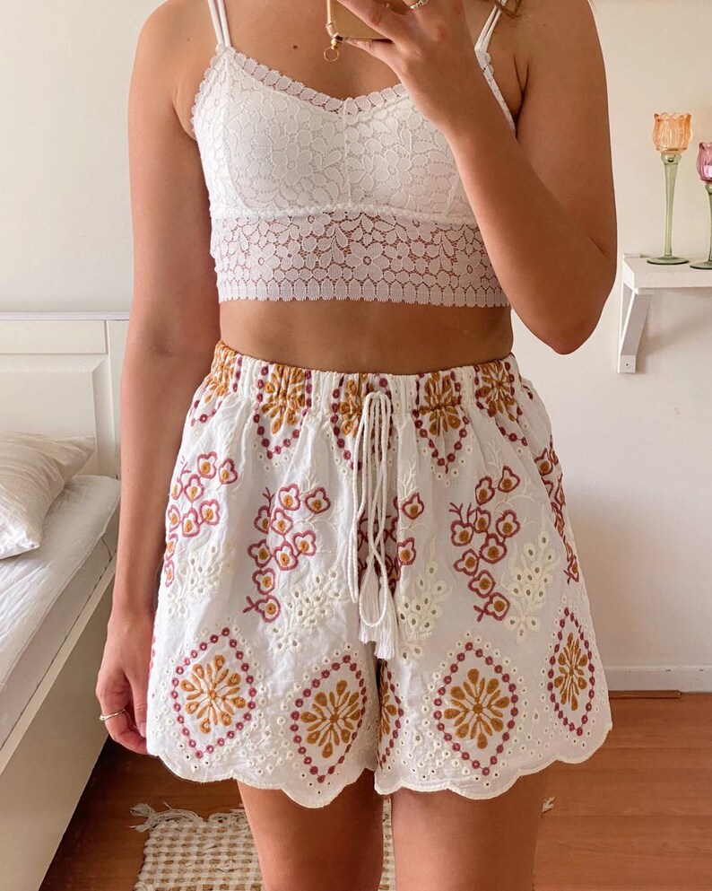 K&ouml;nnte beinhalten: Wei&szlig;e H&auml;kelshorts mit einem gewellten Saum und gestickten Blumen- und geometrischen Mustern in Braun-, Rosa- und Wei&szlig;t&ouml;nen.