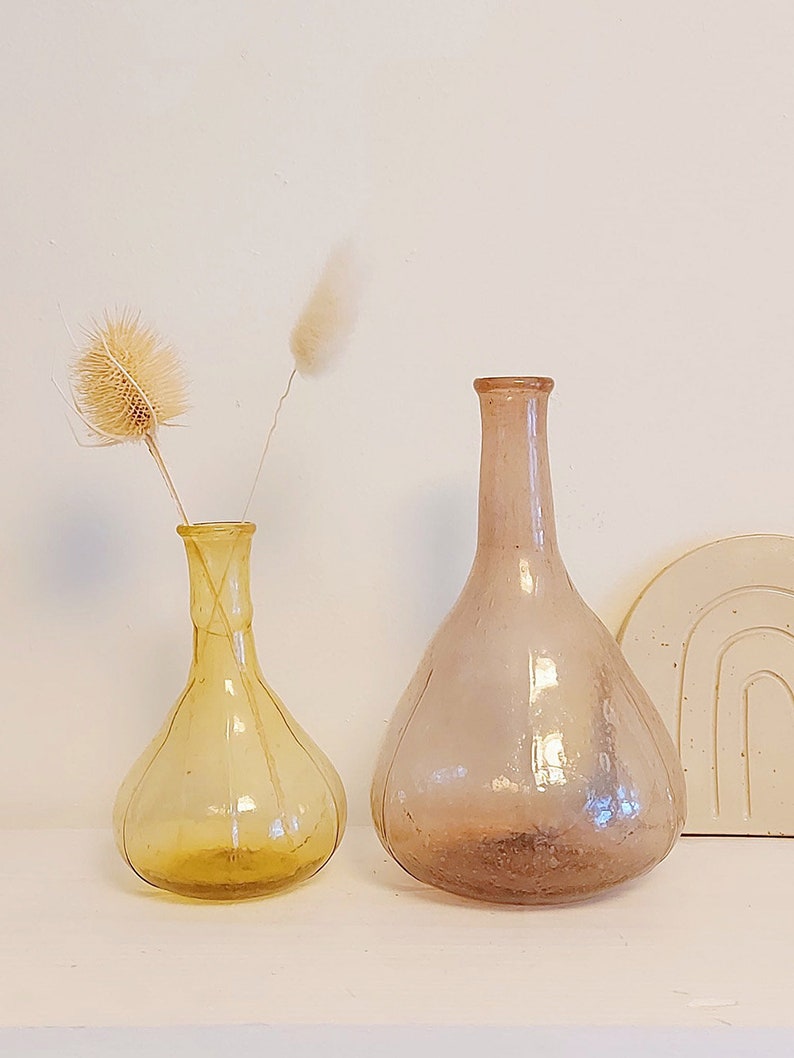 May include: Two glass vases, one amber and one clear with a pink hue. The amber vase has a bulbous shape with a narrow neck and is filled with dried flowers. The clear vase has a wider base and a long neck.