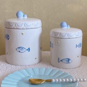 May include: Two white ceramic canisters with blue fish illustrations and light blue lids featuring small spherical knobs. A light blue scalloped plate sits in front with a gold-toned spoon featuring a handle decorated with white beads.