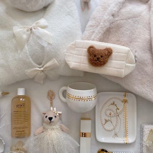 May include: An assortment of white and cream-colored items, including a sweater with bows, a teddy bear keychain, a cosmetic bag with a teddy bear face, a mug with gold accents, and a perfume bottle. The items are arranged on a soft, textured surface.