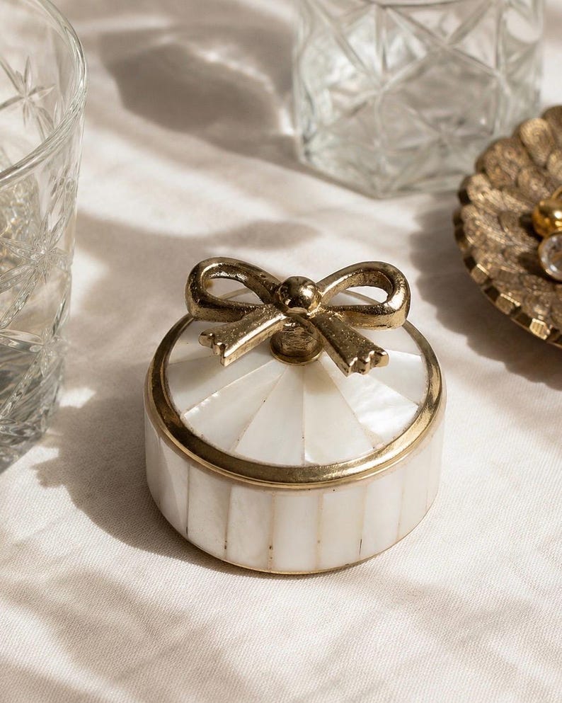 Puede incluir: Un joyero redondo con tapa que presenta una incrustaci&oacute;n de concha blanca iridiscente dispuesta en un patr&oacute;n radial en la tapa y tiras verticales en la base. La caja est&aacute; acentuada con un borde met&aacute;lico dorado y un asa decorativa en forma de lazo.