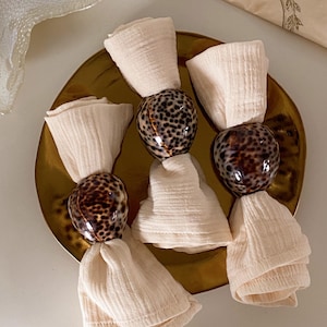 May include: Three cream-coloured cloth napkins are held together by decorative, speckled shell napkin rings. The napkins are arranged on a gold-coloured plate, creating an elegant table setting. The shells have a glossy finish.