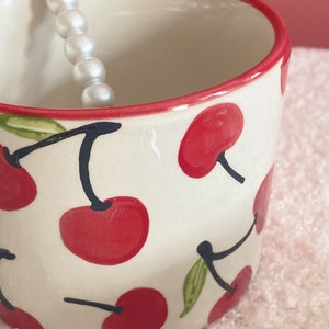 May include: A white ceramic mug with a red rim and a cherry pattern. The mug is decorated with red cherries and green leaves on a white background.